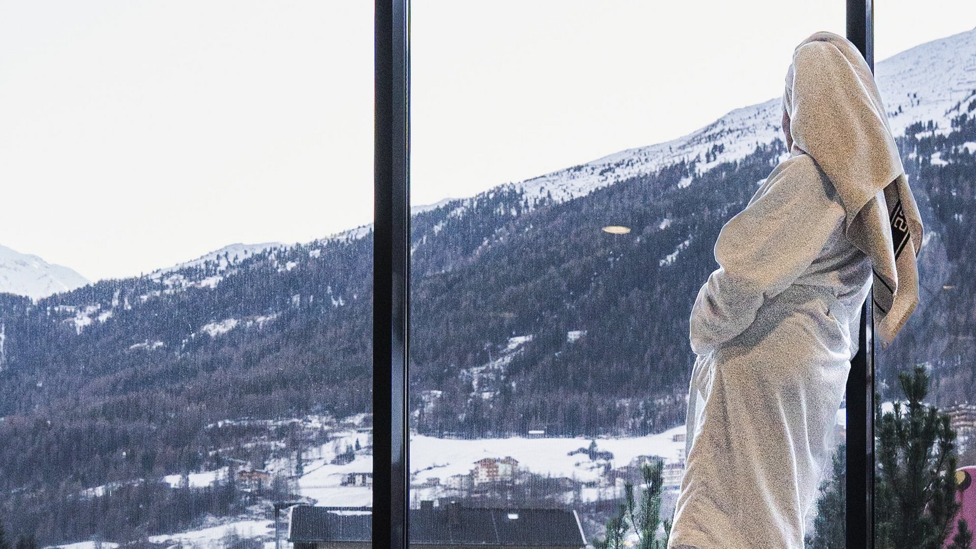 Wellnesshotel in Sölden: Hotel Sunny Person im Bademantel vor großen Fenstern mit verschneiten Bergen und Spiegelung im Wasser