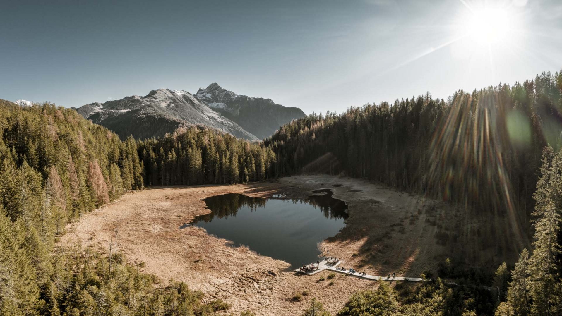 Ausflugsziele in Sölden gesucht? Bergsee mit trockenem Ufer, umgeben von Wald und Bergen im Sonnenlicht