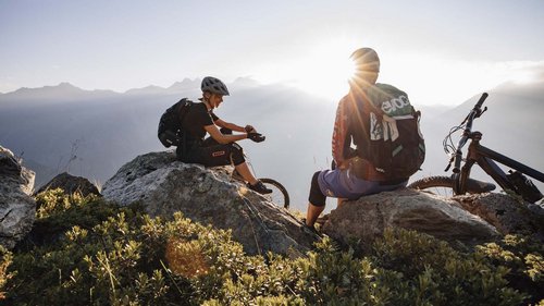 Dein 4-Sterne-Hotel im Ötztal Zwei Mountainbiker machen Pause auf Felsen mit Blick auf Berge und Sonnenuntergang.