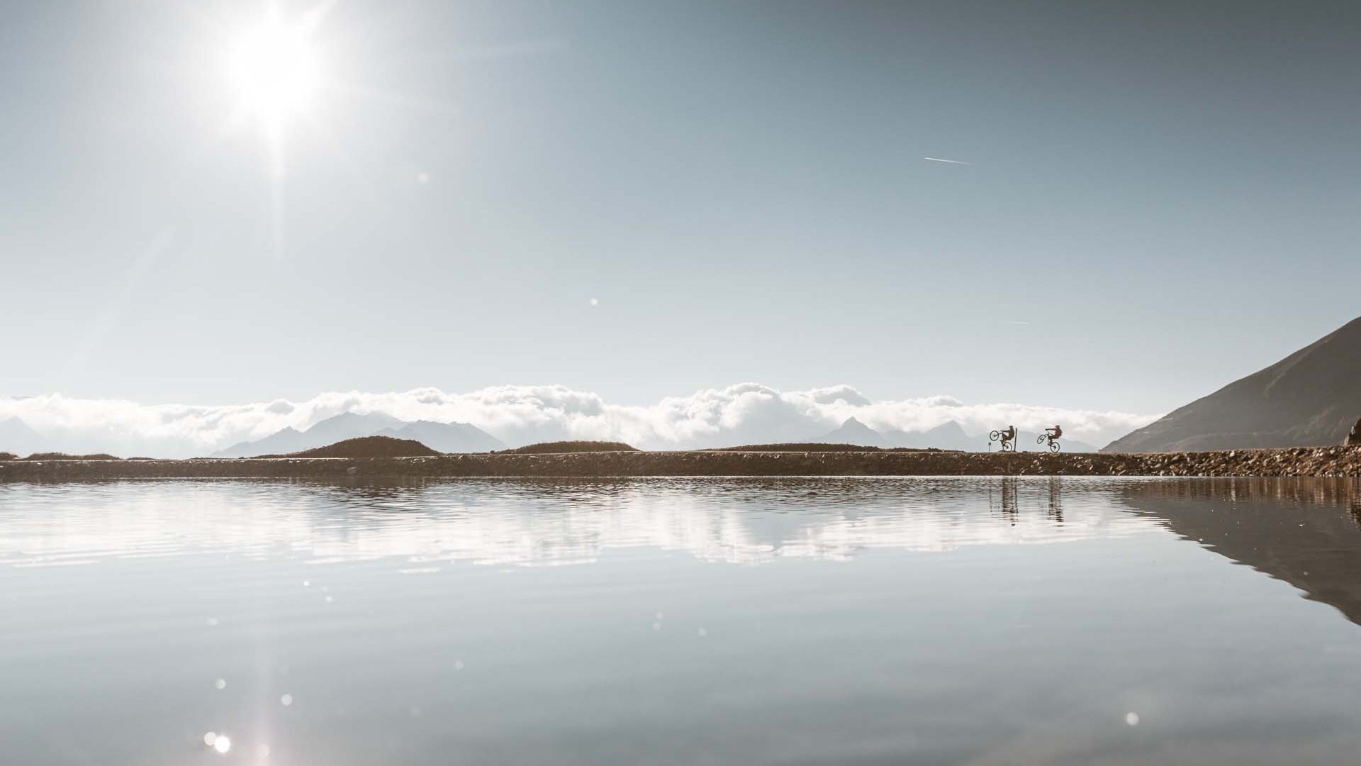 Biking hotel in Sölden: Hotel Sunny Two cyclists doing wheelies by a lake with sunny sky and mountains in background