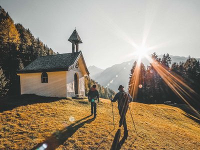 Wandern in Sölden? Im Sommer und Winter! Zwei Wanderer gehen bei Sonnenuntergang an einer kleinen Kapelle auf einem Hügel vorbei