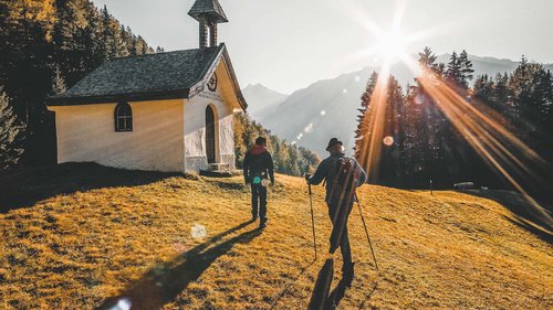 Dein 4-Sterne-Hotel im Ötztal Zwei Wanderer gehen bei Sonnenuntergang an einer kleinen Kapelle auf einem Hügel vorbei