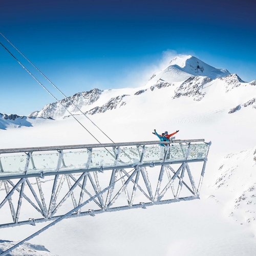 Skiing holiday in Sölden? Come to Hotel Sunny! Bridge in the Alps with two people against snowy mountain backdrop.
